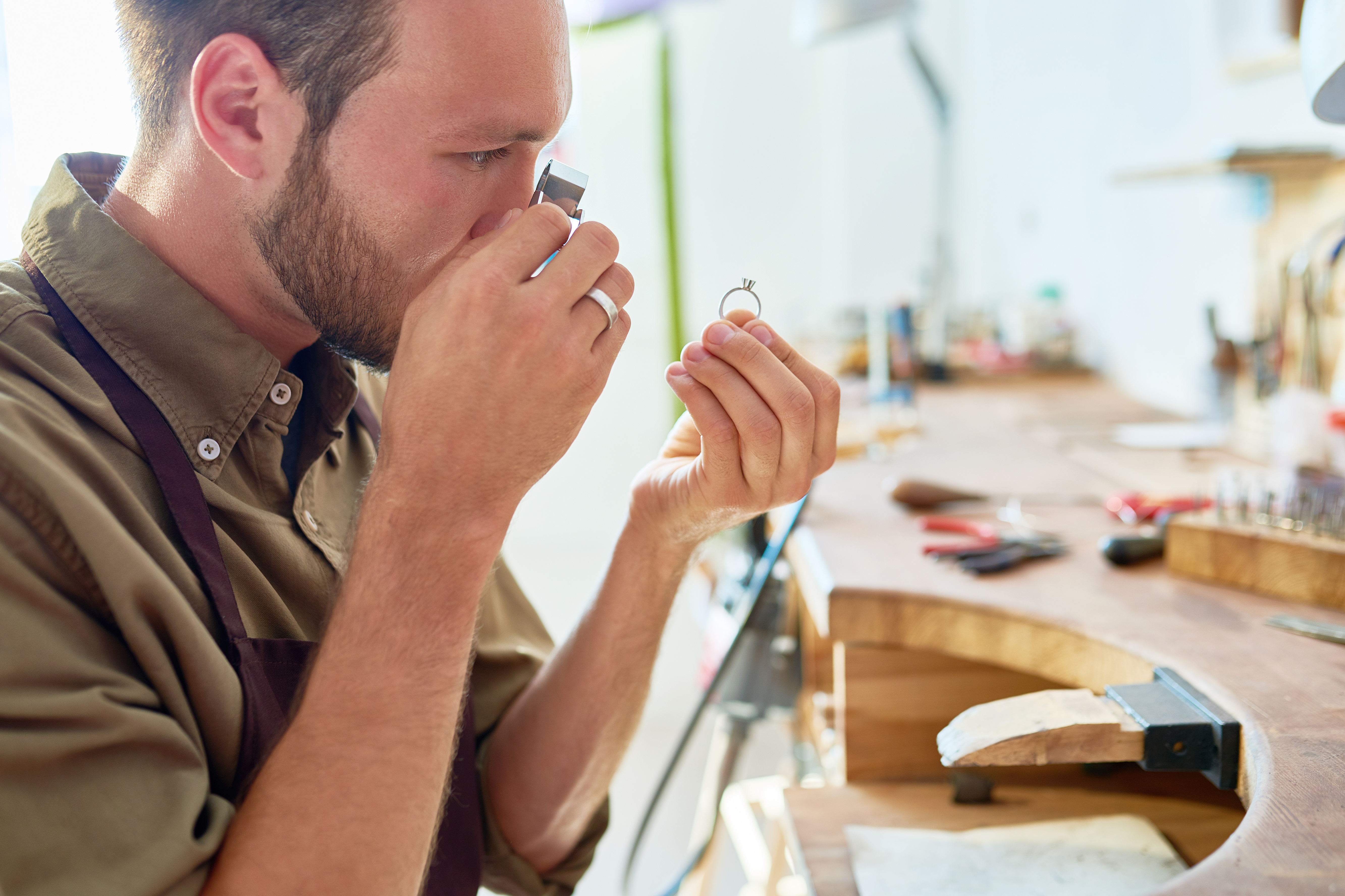 Man inspecting a ring closely using a jeweler’s loupe in a jewelry workshop.