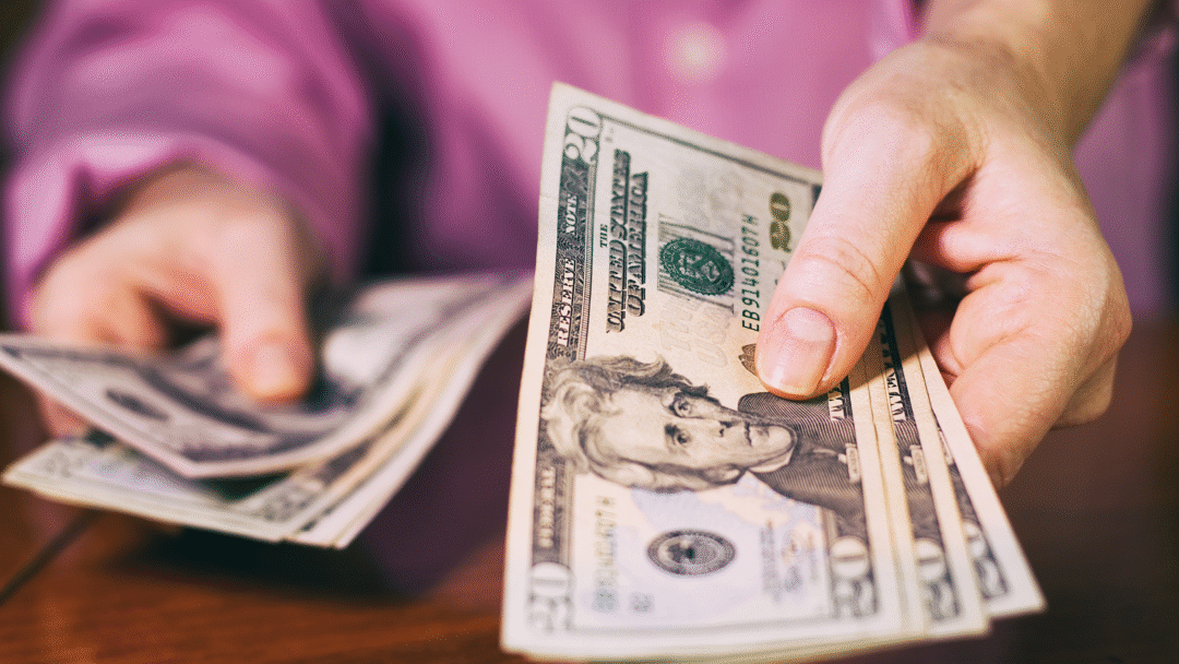 Close-up of a person holding and counting twenty-dollar bills at a table.