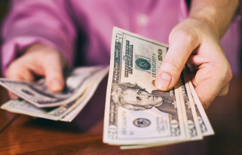 Close-up of a person holding and counting twenty-dollar bills at a table.