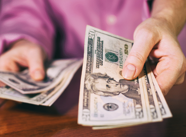 Close-up of a person holding and counting twenty-dollar bills at a table.