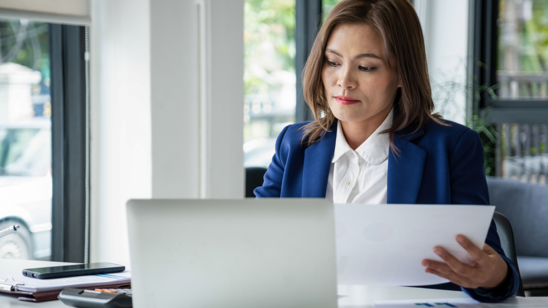 Woman in an office reviewing documents while working on a laptop at her desk