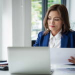 Woman in an office reviewing documents while working on a laptop at her desk