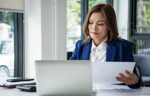 Woman in an office reviewing documents while working on a laptop at her desk