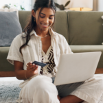 Person sitting on the floor using a laptop while holding a credit card at home.