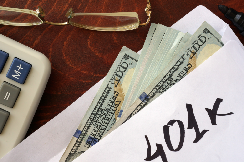 Envelope labeled “401k” with cash partially visible, next to a calculator and eyeglasses on a desk.