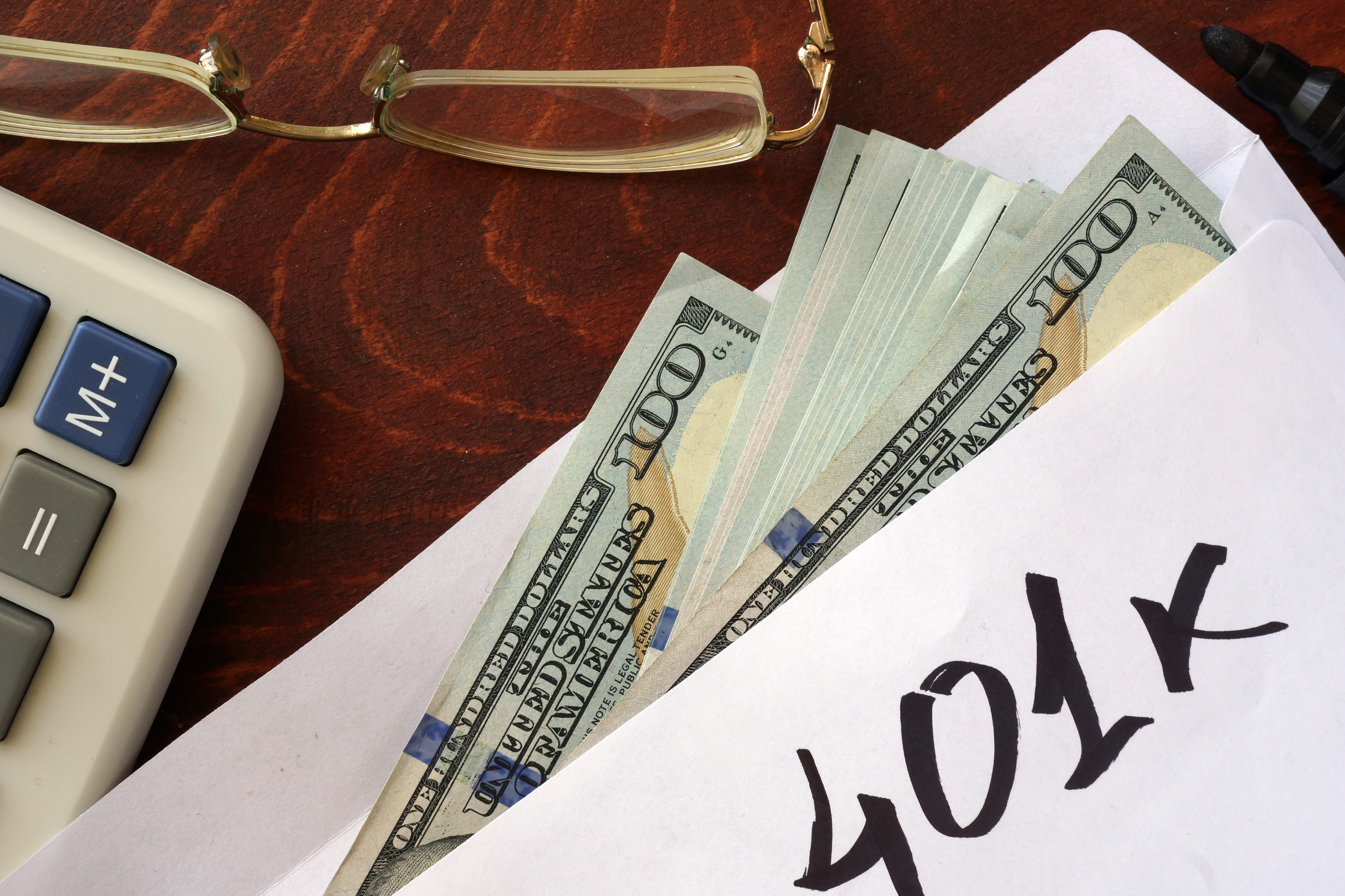 Envelope labeled “401k” with cash partially visible, next to a calculator and eyeglasses on a desk.