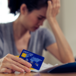 Person holding a credit card while reviewing bills at a table, with their hand on their forehead in a worried pose.