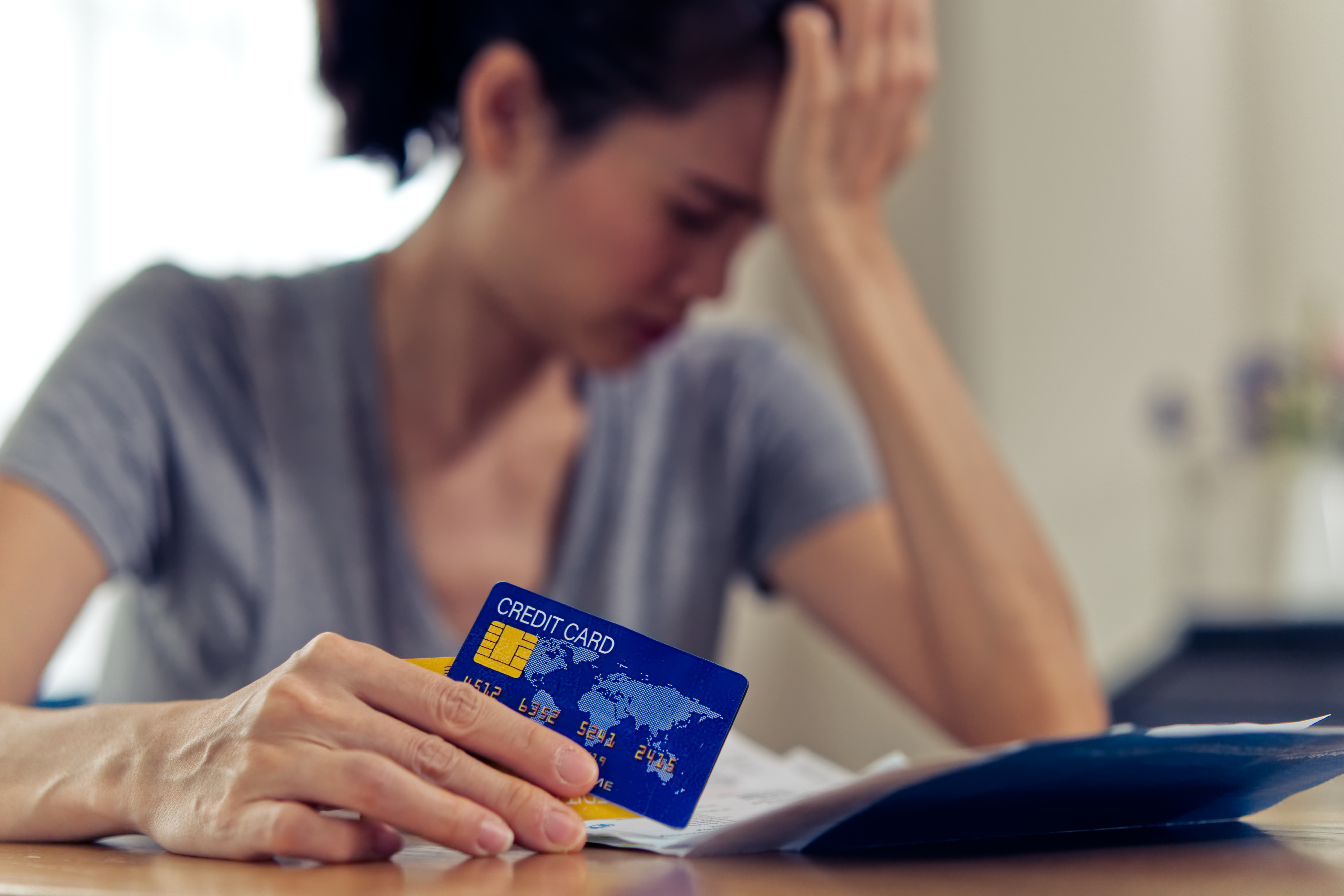 Person holding a credit card while reviewing bills at a table, with their hand on their forehead in a worried pose.