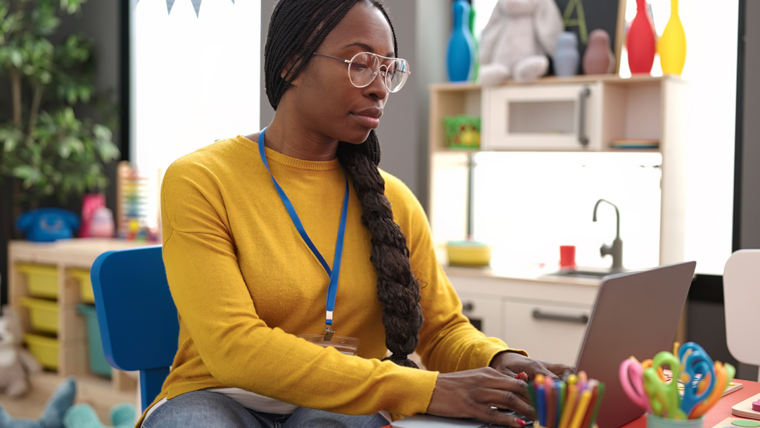 Teacher using a laptop at a small desk in a colorful classroom, with school supplies and toys in the background.