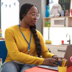 Teacher using a laptop at a small desk in a colorful classroom, with school supplies and toys in the background.