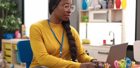 Teacher using a laptop at a small desk in a colorful classroom, with school supplies and toys in the background.