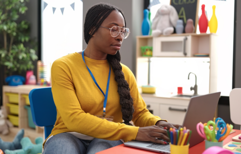Teacher using a laptop at a small desk in a colorful classroom, with school supplies and toys in the background.