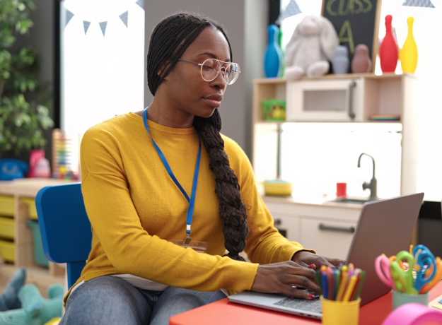 Teacher using a laptop at a small desk in a colorful classroom, with school supplies and toys in the background.
