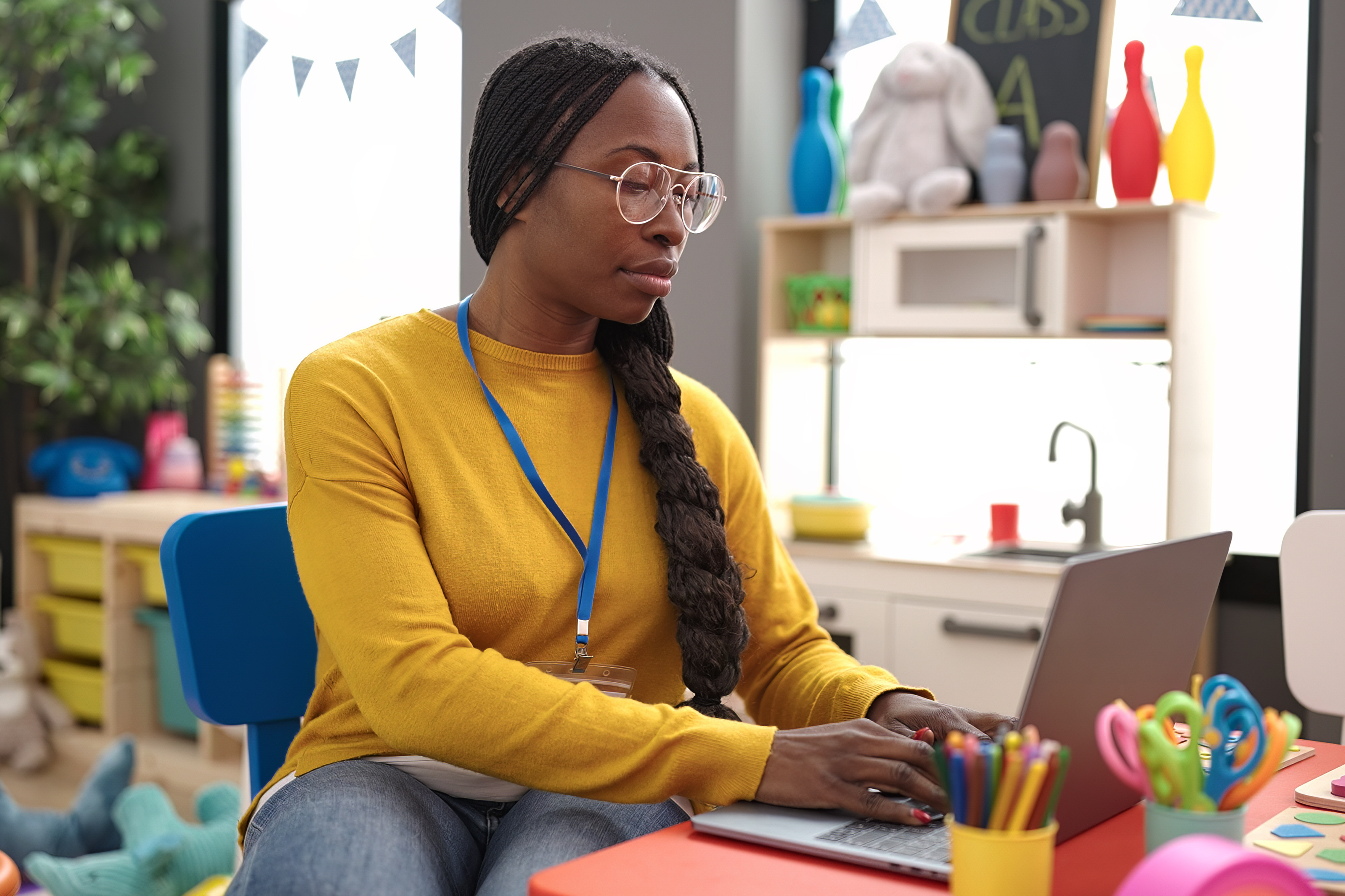 Teacher using a laptop at a small desk in a colorful classroom, with school supplies and toys in the background.