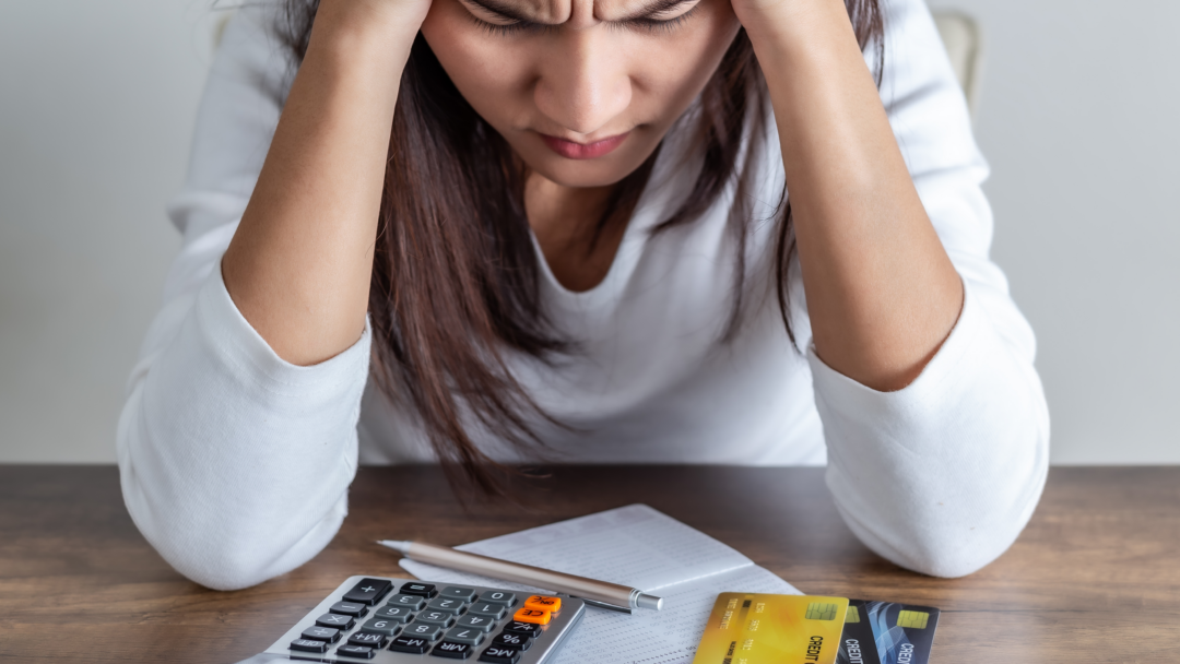 Person holding their head while looking down at a calculator, receipt, and credit cards on a table.
