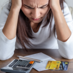 Person holding their head while looking down at a calculator, receipt, and credit cards on a table.