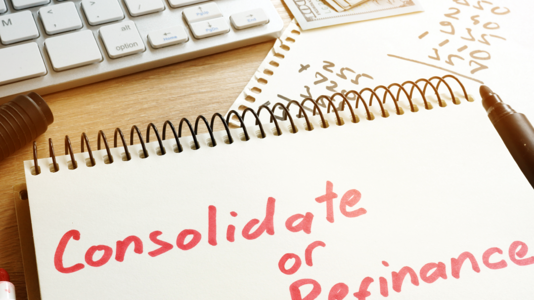 Notebook with the words “Consolidate or Refinance” written in red marker, next to a calculator, keyboard, and cash on a desk.