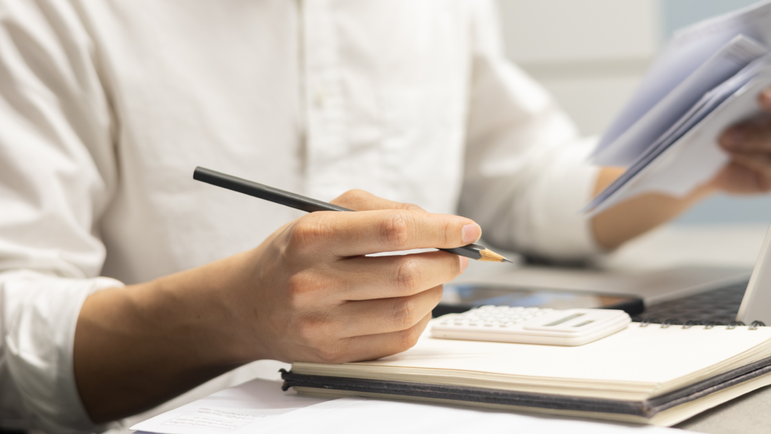 Close-up of a person holding a pencil and reviewing paperwork beside a calculator, notebook, and laptop on a desk.