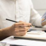Close-up of a person holding a pencil and reviewing paperwork beside a calculator, notebook, and laptop on a desk.