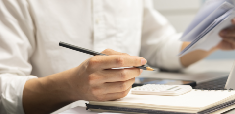 Close-up of a person holding a pencil and reviewing paperwork beside a calculator, notebook, and laptop on a desk.