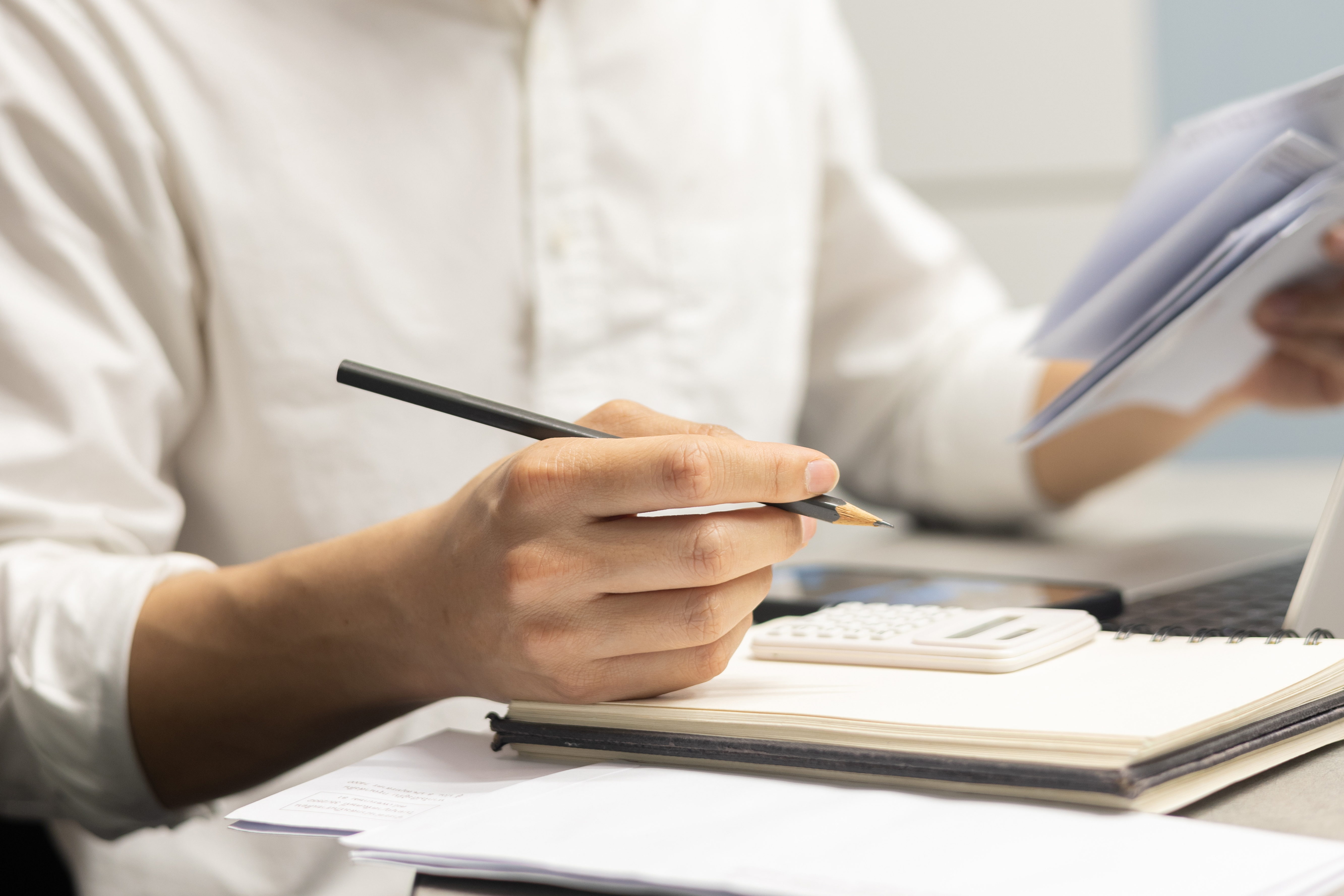 Close-up of a person holding a pencil and reviewing paperwork beside a calculator, notebook, and laptop on a desk.