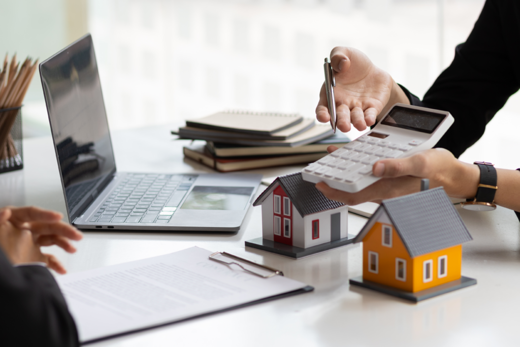 Person using a calculator near model houses and a laptop, suggesting real estate or mortgage planning