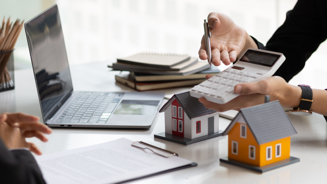 Person using a calculator near model houses and a laptop, suggesting real estate or mortgage planning