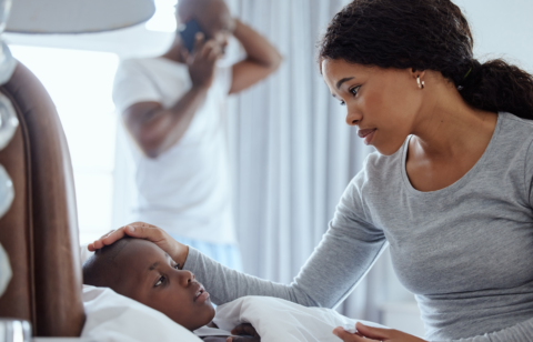 Mother checking on a sick child in bed while a man talks on the phone in the background.