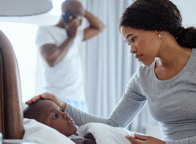 Mother checking on a sick child in bed while a man talks on the phone in the background.