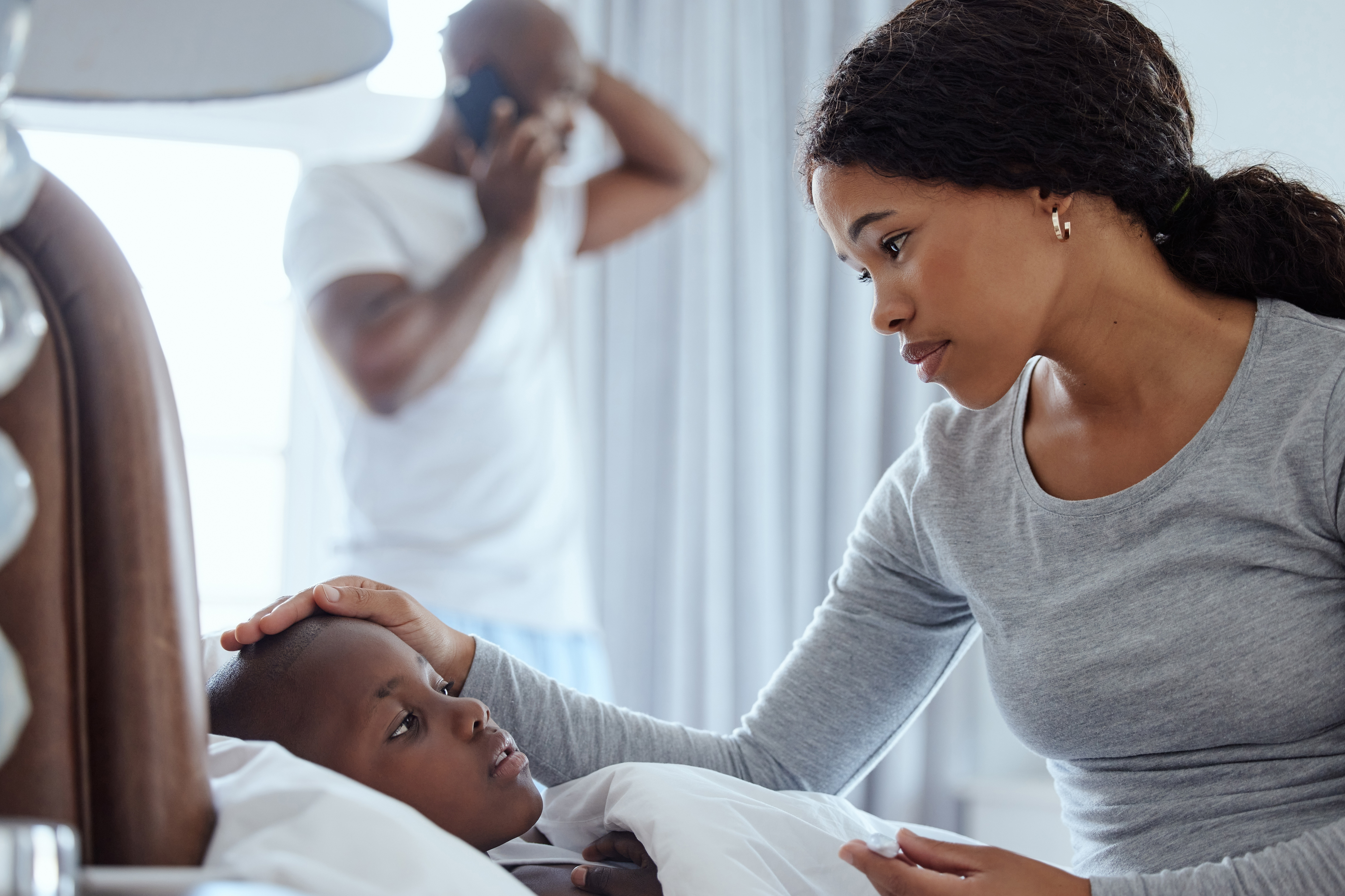 Mother checking on a sick child in bed while a man talks on the phone in the background.