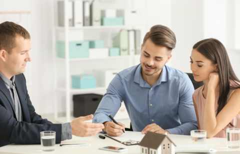 Couple signing paperwork with a professional at a desk, with a small house model in front