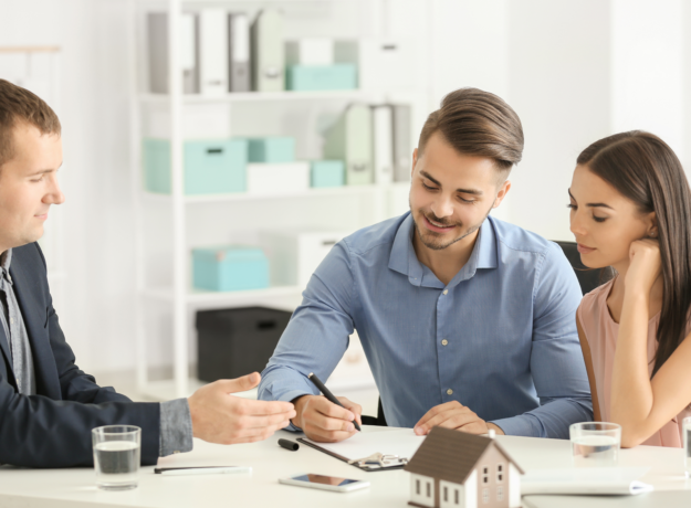 Couple signing paperwork with a professional at a desk, with a small house model in front