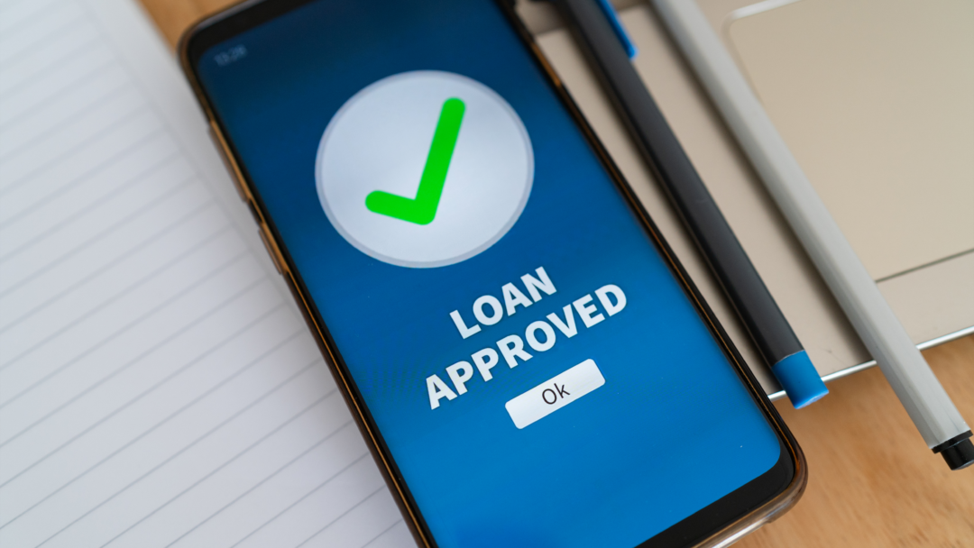 Smartphone screen displaying a green check mark and the words “Loan Approved” next to a notebook and pen.