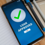 Smartphone screen displaying a green check mark and the words “Loan Approved” next to a notebook and pen.