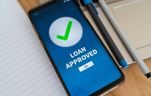 Smartphone screen displaying a green check mark and the words “Loan Approved” next to a notebook and pen.