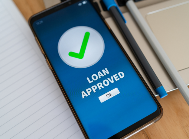 Smartphone screen displaying a green check mark and the words “Loan Approved” next to a notebook and pen.