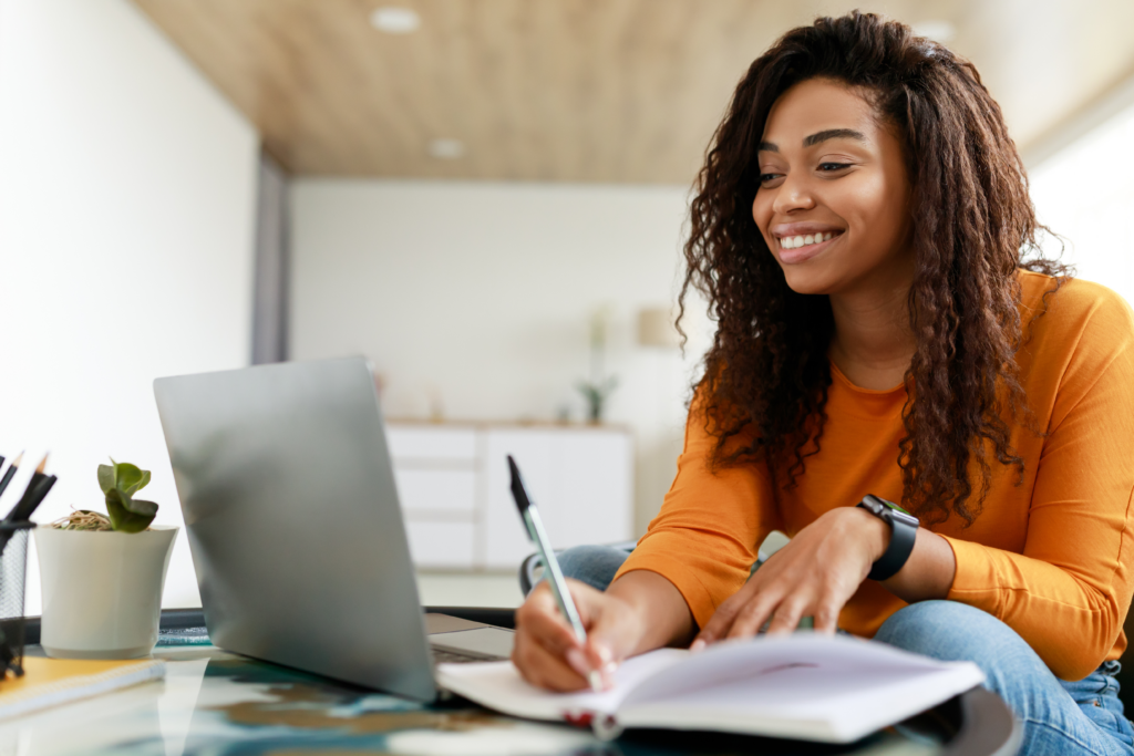 Woman smiling while using a laptop and writing in a notebook at home.