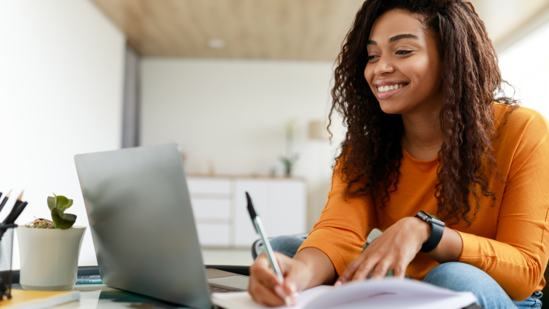 Woman smiling while using a laptop and writing in a notebook at home.