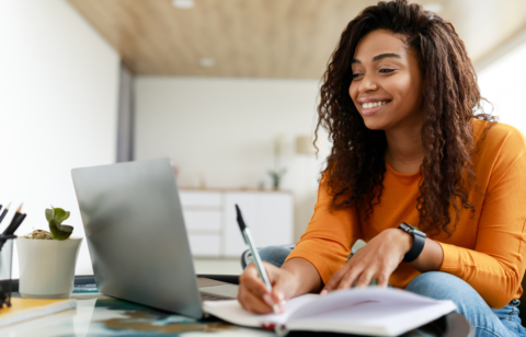 Woman smiling while using a laptop and writing in a notebook at home.