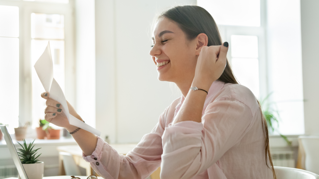 Smiling woman sitting at a desk, holding and reading a letter while looking pleased, with a laptop open in front of her.