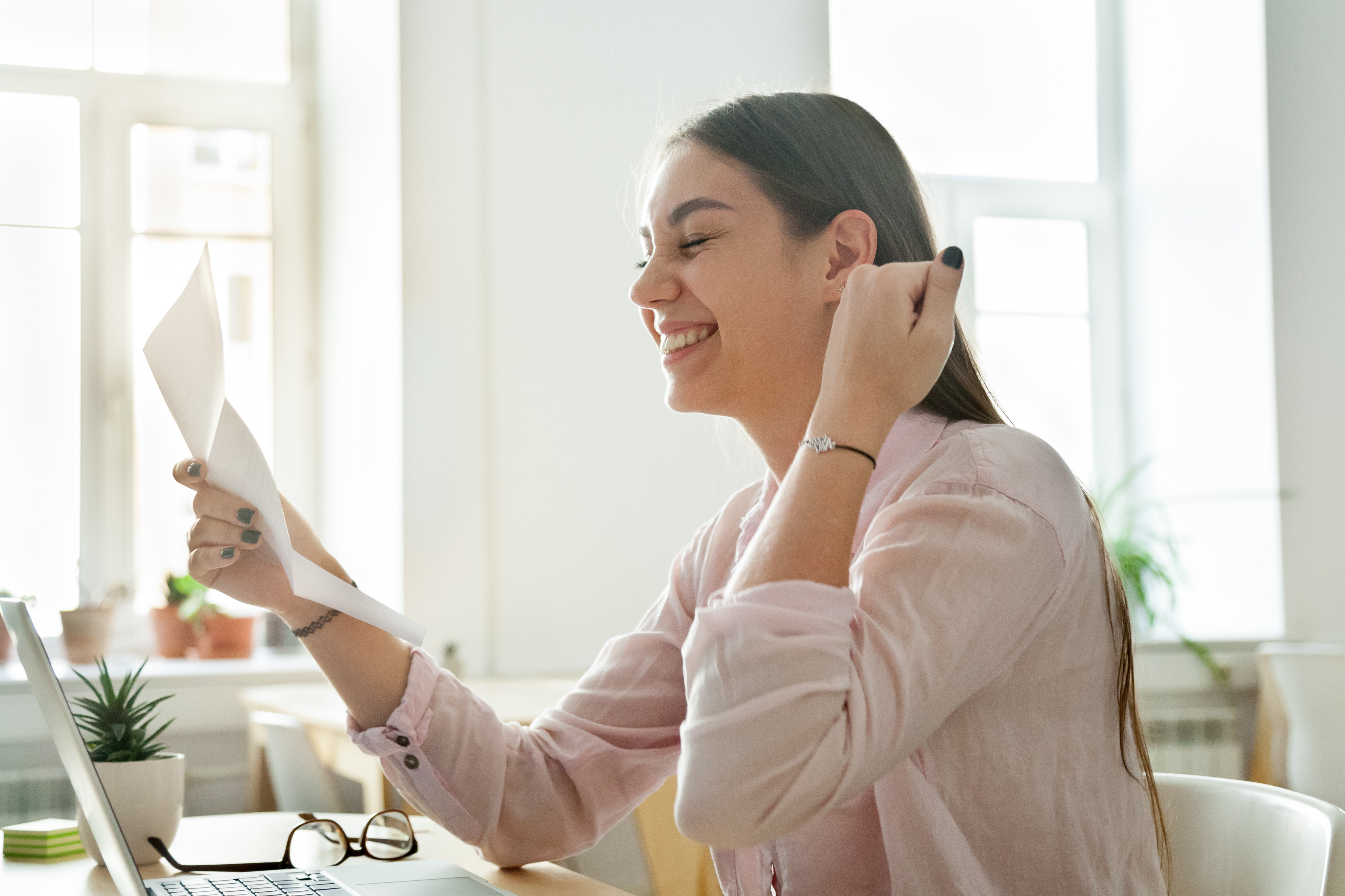 Smiling woman sitting at a desk, holding and reading a letter while looking pleased, with a laptop open in front of her.