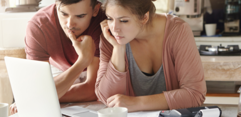 A young couple reviews bills and financial documents on a laptop at their kitchen table, with a calculator and paperwork spread out in front of them.