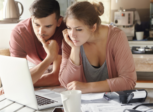 A young couple reviews bills and financial documents on a laptop at their kitchen table, with a calculator and paperwork spread out in front of them.