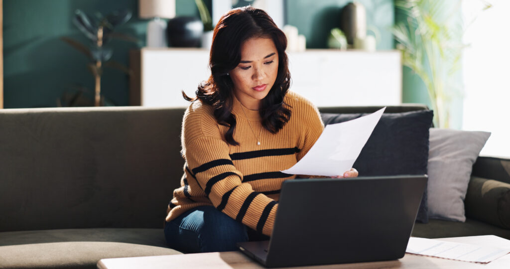 Woman reviewing paperwork and using a laptop while sitting on a couch at home.