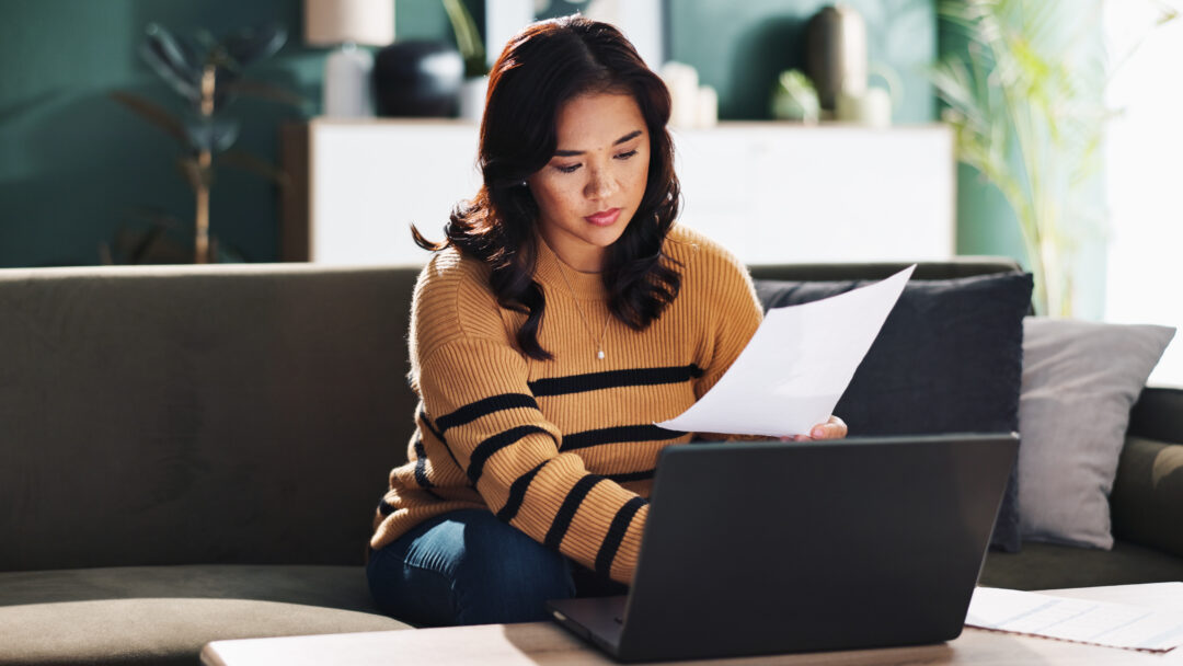 Woman reviewing paperwork and using a laptop while sitting on a couch at home.