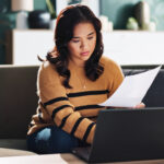 Woman reviewing paperwork and using a laptop while sitting on a couch at home.