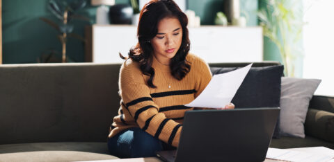 Woman reviewing paperwork and using a laptop while sitting on a couch at home.