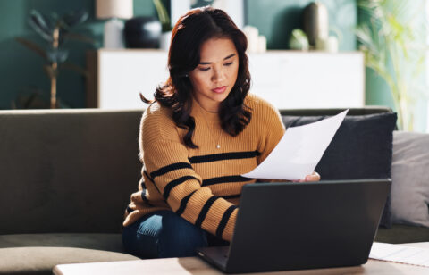 Woman reviewing paperwork and using a laptop while sitting on a couch at home.