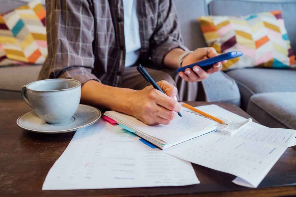 Person writing in a notebook while holding a smartphone, with bills and a cup of coffee on the table.