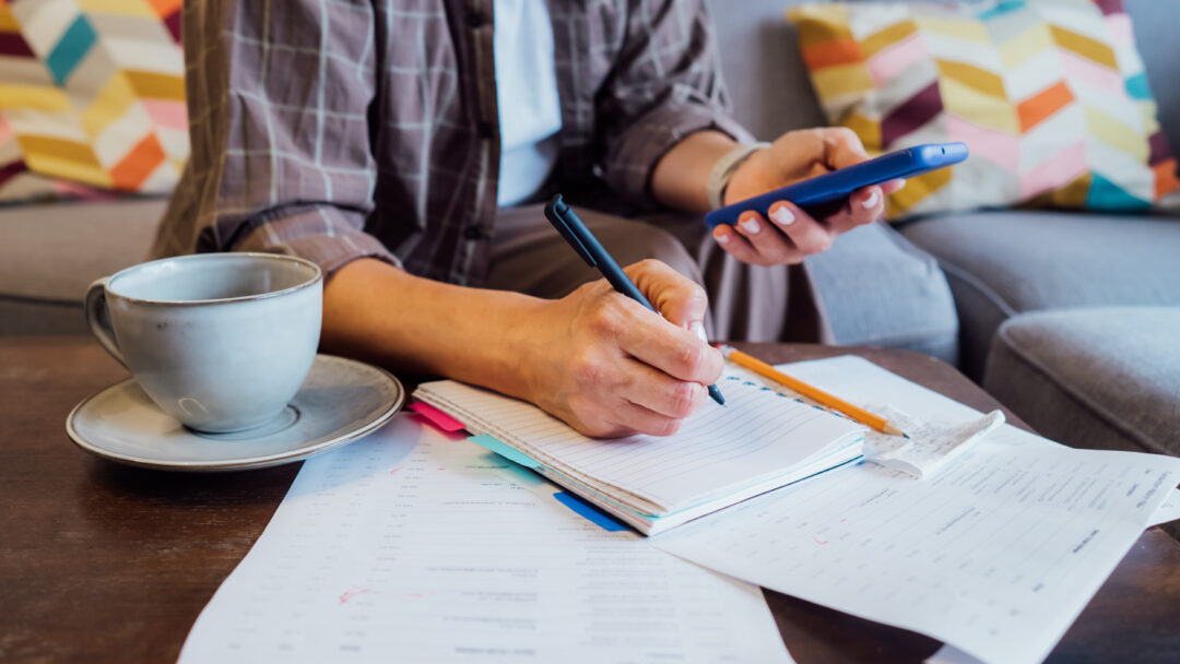 Person writing in a notebook while holding a smartphone, with bills and a cup of coffee on the table.